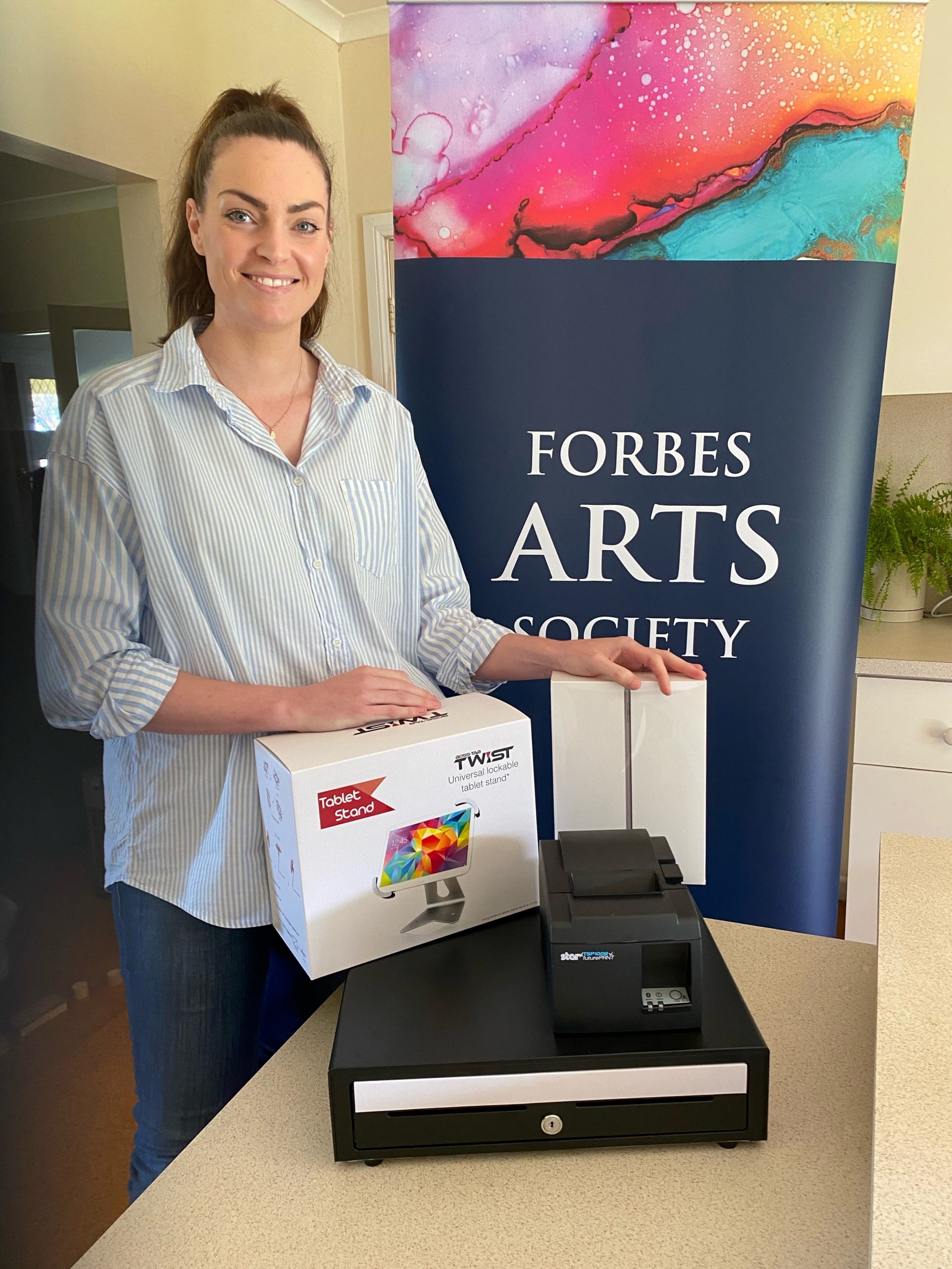 Smiling woman standing in front of "Forbes Arts Society" banner with new point-of-sale equipment on display