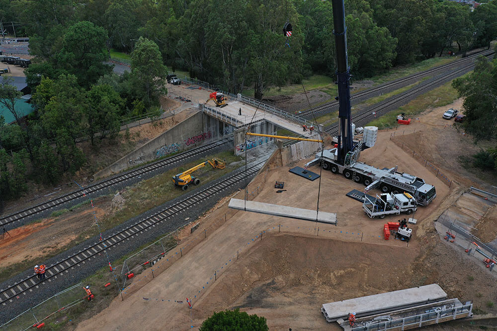 A crane puts a large concrete bridge beam on the ground next to a partially deconstructed bridge.