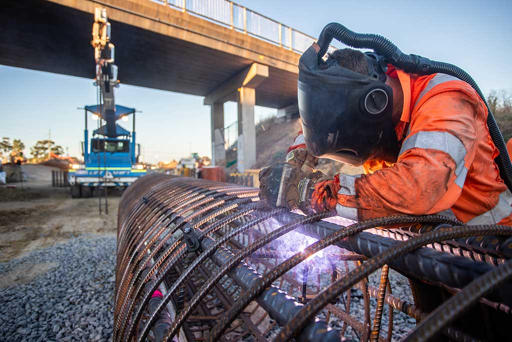 A worker wearing a heavy welding helmet cuts steel mesh at a rail worksite