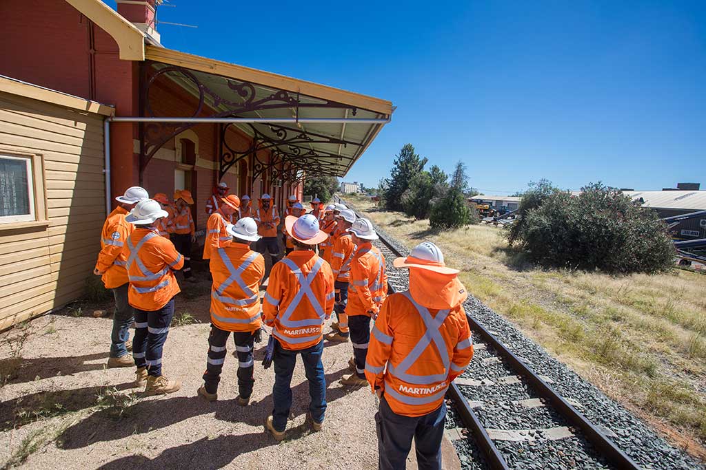 A group of workers wearing orange jackets and hard hats standing under an awning at Forbes Station